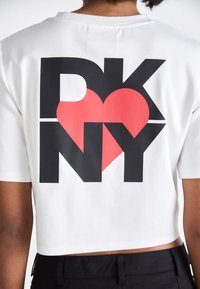 Bílý zkrácený tričko s velkým grafickým potiskem "DKNY," červeným srdcem a "NY" v černé barvě. Materiál vypadá hladce a ležérně.