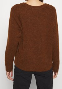 Pull marron en tissu tricoté doux et texturé. Il présente un col rond, des manches longues et un ourlet arrière légèrement arrondi.