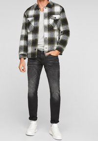 Chaqueta de algodón a cuadros en verde, negro y blanco con dos bolsillos en el pecho, combinada con jeans gris oscuro y zapatillas blancas.