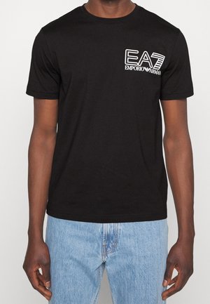 Mann trägt schwarzes EA7 Emporio Armani T-Shirt und hellblaue Jeans, steht vor einem schlichten hellen Hintergrund.