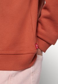 Gros plan d'une personne portant un sweat-shirt orange brûlé avec une étiquette rouge et un pantalon rose clair, la main dans la poche.