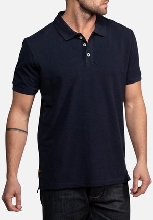 Homme portant un polo bleu marine foncé uni à manches courtes avec trois boutons, assorti à un jean en denim foncé.