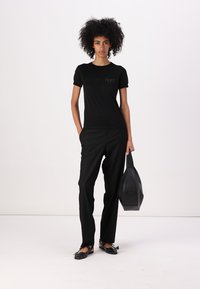 Maglietta nera a maniche corte con logo, abbinata a pantaloni neri e scarpe flat. Con una borsa nera, la modella è in piedi contro uno sfondo bianco.
