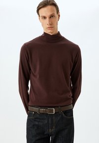 Brauner Rollkragenpullover aus weichem Strickstoff, mit einer körpernahen Passform und langen Ärmeln, kombiniert mit dunklen Jeans und einem braunen Gürtel.