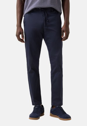 Mand iført marineblå slimfit bukser med snøre i taljen og mørkeblå sneakers med lysebrune såler, stående mod hvid baggrund.