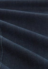 Tumma farkkukangas, jossa on sileä pinta, hienovaraisia sävyeroja ja hieman kiiltävä pinta. Taitetut kerrokset luovat dynaamisen ilmeen.