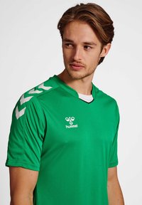 Grünes synthetisches Sporttrikot mit V-Ausschnitt, kurzen Ärmeln und weißen Chevron-Mustern auf den Schultern. Das "Hummel"-Logo ist deutlich sichtbar.