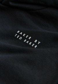 Tissu en coton noir avec un texte blanc indiquant "BAKER BY TED BAKER", positionné de manière bien visible sur un fond texturé.