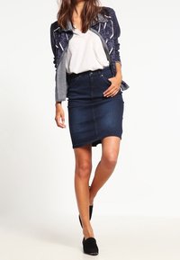 Jeanskjol, mörkblå med en figursydd silhuett, bärs med en ljus t-shirt och mönstrad jacka. Svarta slip-on skor fulländar outfiten.