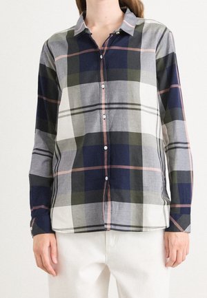 Femme portant une chemise à carreaux à manches longues boutonnée en bleu marine, olive, gris et blanc avec des accents rose clair, assortie à un pantalon blanc.