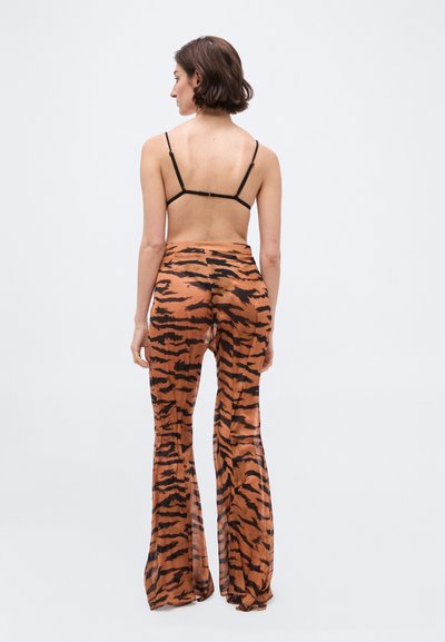 Γυναίκα με την πλάτη γυρισμένη, φορούν λεπτό ραντάκι μαύρο σουτιέν και φαρδιά παντελόνια με τύπωμα τίγρης και ψηλή μέση, σε απλό φόντο.