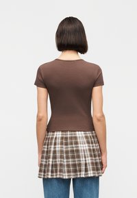 Γυναίκα με κοντά σκούρα μαλλιά π ου φοράει ένα εφαρμοστό καφέ κοντομάνικο πάνω από μια πλισέ καφέ και λευκή καρό φούστα, στρωμένη με μπλε τζιν, γυρισμένη πλάτη.