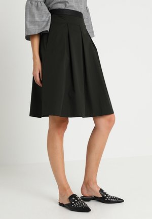 Kvinde iført sort knælange plisseret nederdel, grå ternet top med pufærmer og sorte spidse slip-on sko med sølvnitter.