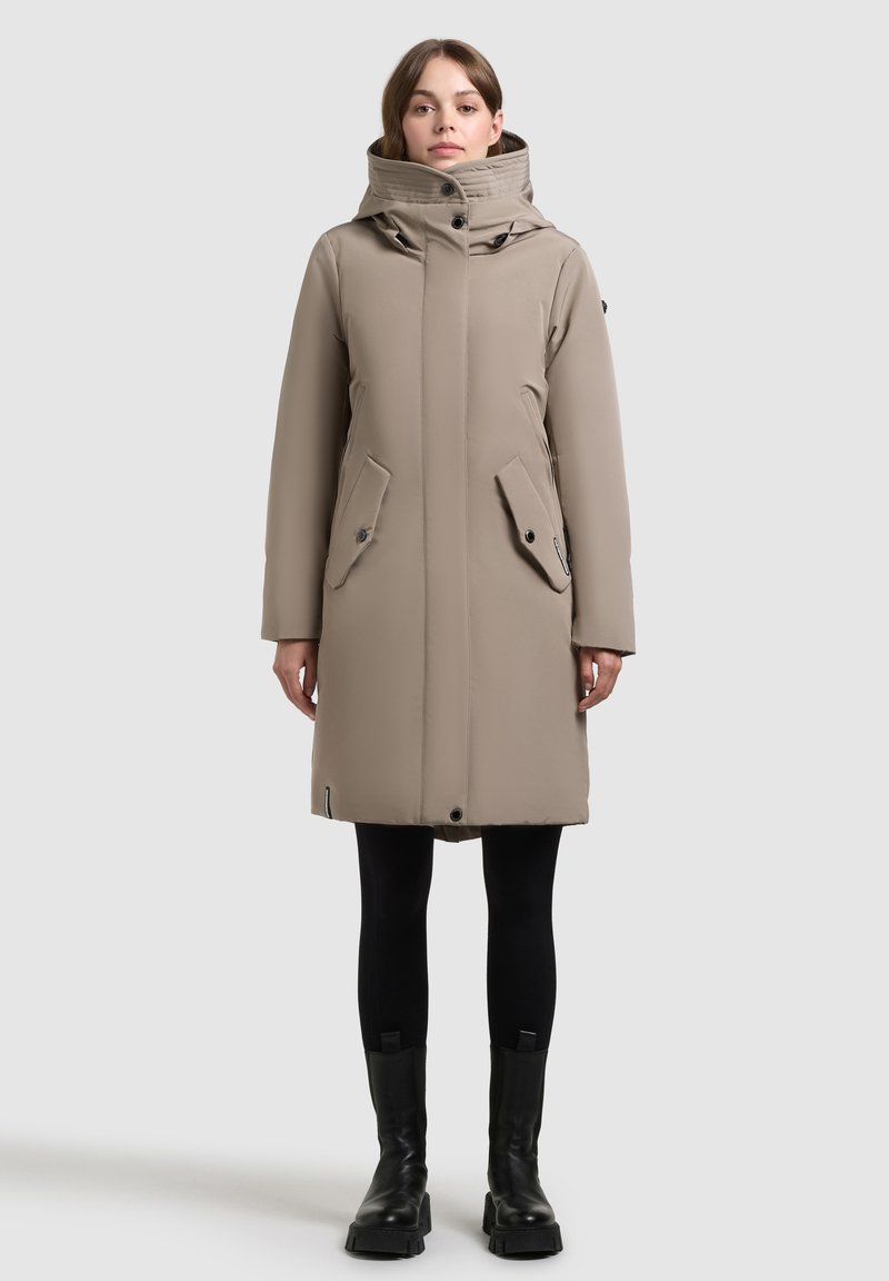 Beige mid-length parka with a high collar, double snap closures, side pockets, and a smooth, textured finish. Worn over black leggings and boots.