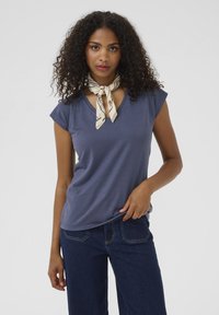 Kortärmad, mörkblå V-ringad t-shirt i mjukt tyg, matchad med mörka jeans. En mönstrad ljus scarf är knuten runt halsen.