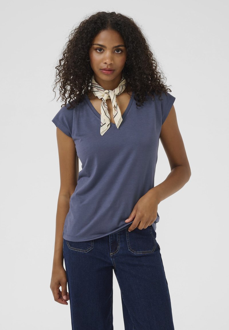 Kortärmad, mörkblå V-ringad t-shirt i mjukt tyg, matchad med mörka jeans. En mönstrad ljus scarf är knuten runt halsen.