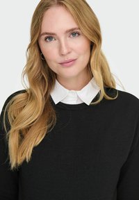 Musthave musta sviiter ümmarguse kaelusega, all valge kraega särgiga. Sile tekstuur ja lihtne disain. Pikad lainelised blondid juuksed.