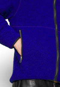 Futrzana kurtka na zamek w żywym niebieskim kolorze o gładkiej fakturze. Posiada kontrastujące czarne mankiety oraz dwie przednie kieszenie na zamek.