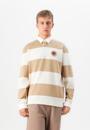 Young man wearing beige and white striped long sleeve polo shirt and light brown pants, standing against plain white background.