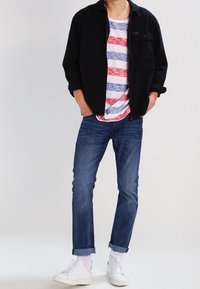 Svart skjortjacka över en röd och blårandig T-shirt, matchad med uppvikta blå jeans och vita sneakers. Enkelt design, ledig passform.