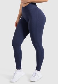 Granatowe legginsy z wysokim stanem, z teksturowanym pasem, bezszwowym designem i detalami na bokach; zestawione z białymi butami sportowymi.