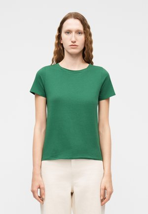 Femme aux cheveux roux ondulés portant un t-shirt vert à manches courtes et un col rond, et un pantalon beige, debout devant un fond clair uni.