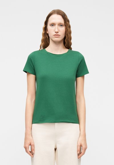 Femme aux cheveux roux ondulés portant un t-shirt vert à manches courtes et un col rond, et un pantalon beige, debout devant un fond clair uni.