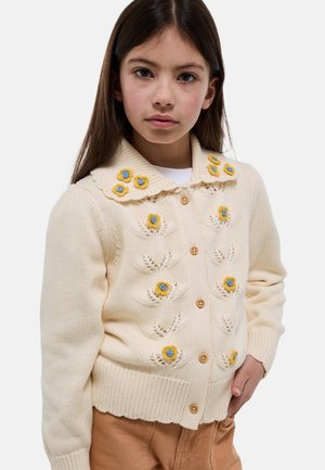 Jeune fille portant un cardigan en tricot crème avec des motifs floraux jaunes et bleus, debout avec la main dans la poche sur un fond blanc.