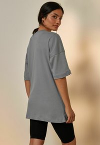 Szary, oversize t-shirt z krótkimi rękawami i zawiniętymi mankietami, gładka faktura, noszony na czarnych leginsach, pokazany z lekko skośnego tylnego widoku.