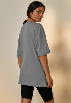 Grijze oversized t-shirt met korte mouwen en opgerolde mouwen, gladde textuur, gedragen over zwarte leggins, getoond vanuit een iets schuin achteraanzicht.