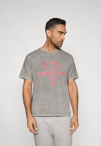 Šedé bavlněné tričko s červeným nápisem "U.S. MEN'S NATIONAL SOCCER TEAM 2026", kulatým výstřihem a krátkými rukávy.