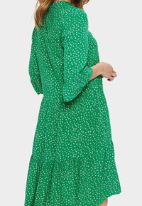 Zelené šaty s vrstevnatým designem, které mají vzor malých bílých teček. Dlouhé rukávy a kulatý výstřih, vyrobené z lehkého materiálu.