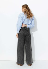 Chemisier bleu clair à rayures avec broderies florales, associé à un jean noir taille haute et large, et des chaussures à motifs léopard, vu de derrière.