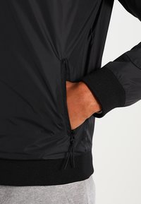 Černá bunda s kapsou na zip, elastickými manžetami a hladkým, lehkým materiálem. Design zahrnuje stahovací šňůrku na lemu.