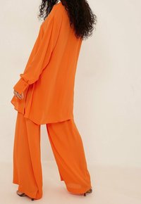 Blouse orange, coupe lâche avec des manches longues, associée à un pantalon large en tissu et couleur assortis, avec une texture lisse et légère.