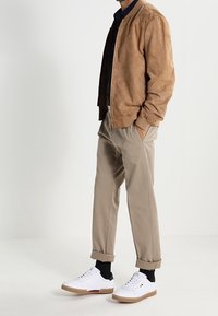 Ruskea suede-bomber-takki, tummapaitainen, vaaleanbeige-housut ja valkoiset tennarit ruskeilla pohjilla. Käännetyt hihansuut, minimalistinen muotoilu.