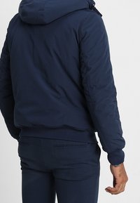 Person med en marinblå huva jacka och matchande byxor, ståendes med ryggen mot kameran mot en ljus bakgrund.