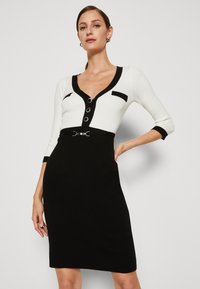 Robe noir et blanc avec un design ajusté, texture côtelée, décolleté en v boutonné et ceinture accentuée à la taille. Manches trois-quarts.