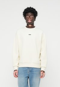 Sudadera de cuello redondo en color blanco roto, con puños y dobladillo acanalados. Presenta un pequeño logotipo negro "BOSS" en el pecho. Combinada con jeans azules.