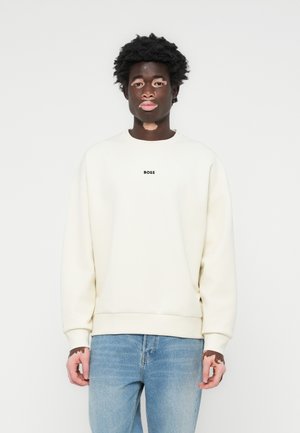 Crewneck sweatshirt in off-white, with ribbed cuffs and hem. Features small black "BOSS" logo on the chest. Paired with blue jeans.