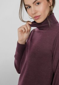Suéter de cuello alto morado con un collar texturizado, mangas ajustadas y un acabado suave, usado por una persona que sostiene el cuello.