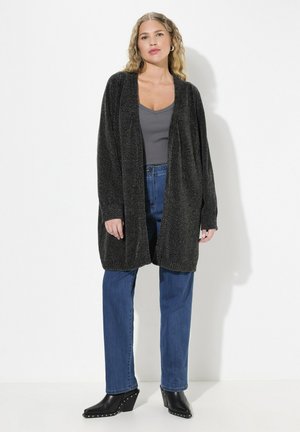 Largo cárdigan negro de apertura frontal con un acabado texturizado, llevado sobre una blusa gris y jeans azules, combinado con botas negras de cuña con tachuelas.