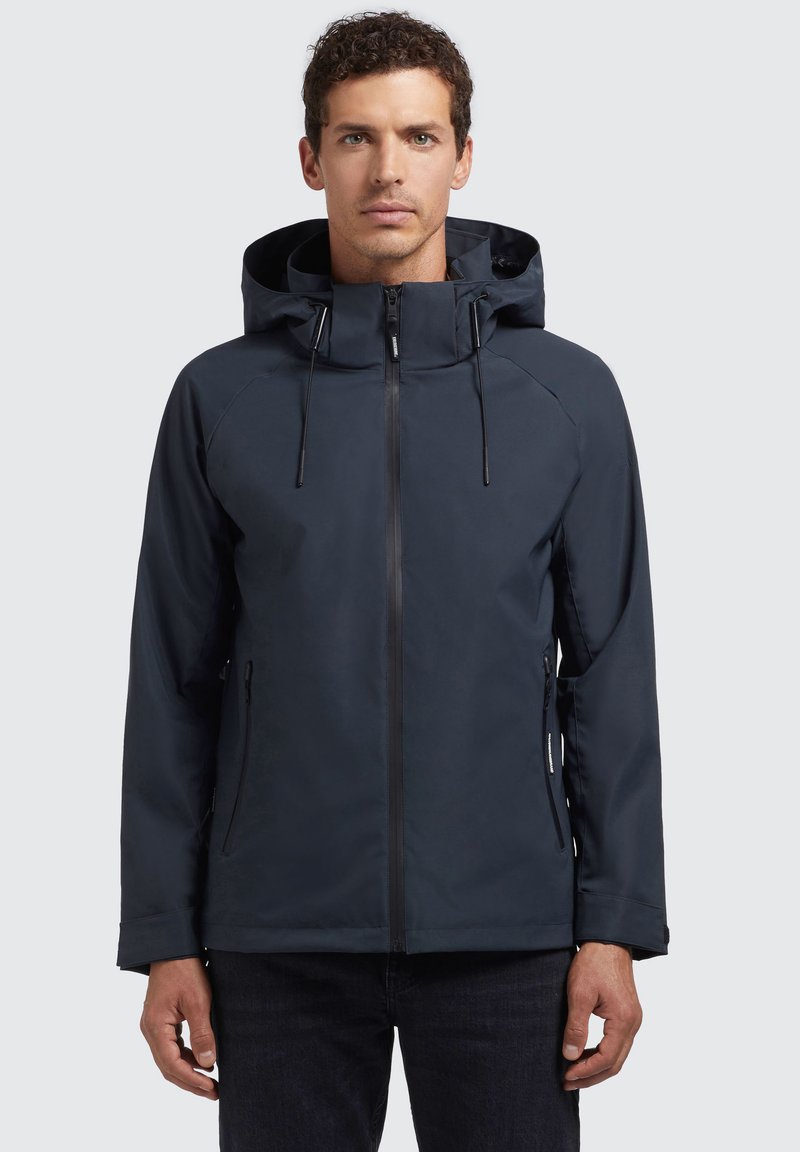 Chaqueta impermeable de color navy con capucha, cremallera frontal vertical, dos bolsillos laterales y cordones ajustables en el cuello. Textura suave y mate.