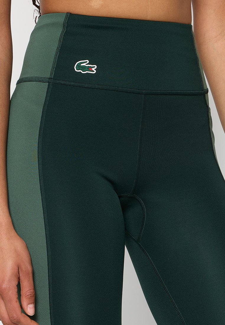 Leggings taille haute en matériau vert foncé avec une texture lisse, présentant un panneau vert contrastant et un logo Lacoste sur la taille.