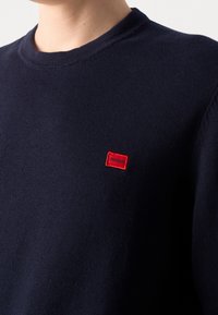 Granatowy sweter z okrągłym dekoltem z małą czerwoną prostokątną łatą z logo "HUGO" na lewej piersi.