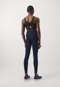 Fekete hálós sportmelltartó megkötő hátrésze. Tengerészkék leggings, testhezálló kialakítással és varrás nélküli derékkal. Sötét sportcipők fehér talppal.