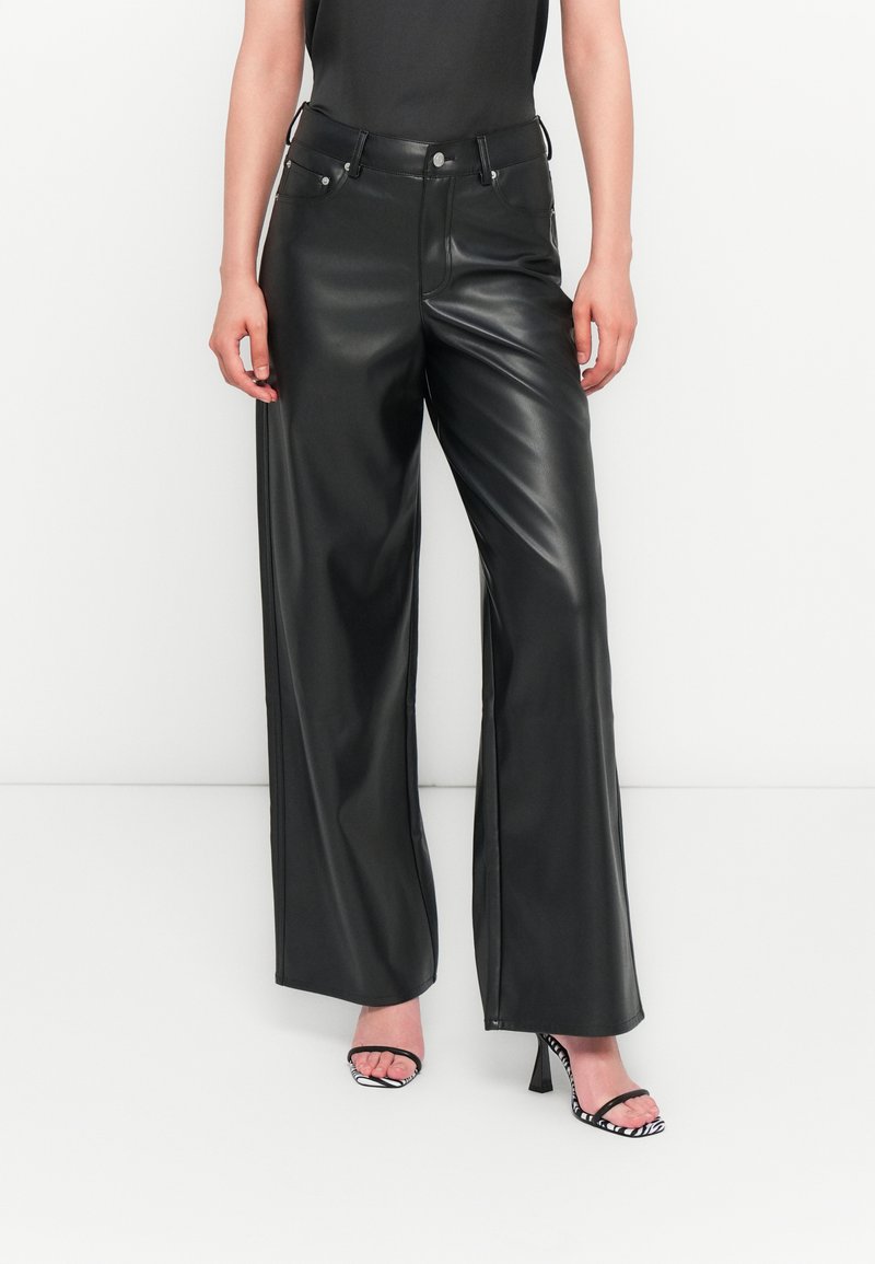 Pantalons larges en simili cuir noir avec taille haute, dotés de poches avant et de quincaillerie en argent, associés à des sandales à talons noires et blanches.