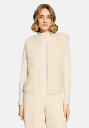 Femme aux cheveux courts, blonds et ondulés, portant un pull à col roulé crème, une veste pelucheuse crème et un pantalon large crème, debout devant un fond clair.