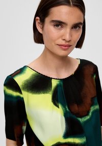 Blouse à manches courtes en tissu lisse. Présente un motif abstrait coloré avec des teintes vertes, jaunes, noires et brunes. Détail plissé au niveau du col.