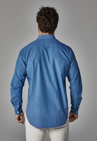 Camisa de manga larga azul clara con puños abotonados, cuello puntiagudo y dobladillo curvado, hecha de una tela suave con un diseño ligeramente estructurado.